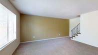 This two-floor space offers a straightforward and functional layout designed to maximize livability across both levels. The main living area on the ground floor is spacious and open, featuring plush wall-to-wall carpeting in a neutral beige tone that adds warmth and comfort underfoot. The walls are finished in a rich golden-tan hue, lending the space a warm, inviting ambiance. Clean white trim and baseboards provide a crisp contrast against the warm wall color, contributing to a tidy and well-defined aesthetic. A staircase positioned toward one side of the living area connects the two floors, finished with a classic wrought iron railing that adds a subtle decorative element to the otherwise understated interior. The ceiling is smooth and white, helping to reflect light and maintain a sense of openness throughout the room. The overall design is clean and minimal, providing a neutral canvas well-suited for a variety of interior styles and furnishing preferences. The second floor expands the available living space, offering additional rooms accessible via the staircase. The combination of warm tones, soft flooring, and classic detailing gives this two-floor space a comfortable and welcoming character throughout.