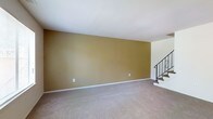 This two-floor space offers a straightforward and functional layout designed to maximize livability across both levels. The main living area on the ground floor is spacious and open, featuring plush wall-to-wall carpeting in a neutral beige tone that adds warmth and comfort underfoot. The walls are finished in a rich golden-tan hue, lending the space a warm, inviting ambiance. Clean white trim and baseboards provide a crisp contrast against the warm wall color, contributing to a tidy and well-defined aesthetic. A staircase positioned toward one side of the living area connects the two floors, finished with a classic wrought iron railing that adds a subtle decorative element to the otherwise understated interior. The ceiling is smooth and white, helping to reflect light and maintain a sense of openness throughout the room. The overall design is clean and minimal, providing a neutral canvas well-suited for a variety of interior styles and furnishing preferences. The second floor expands the available living space, offering additional rooms accessible via the staircase. The combination of warm tones, soft flooring, and classic detailing gives this two-floor space a comfortable and welcoming character throughout.