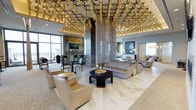 This elegantly designed single-floor space exudes refined luxury through its thoughtful architectural details and curated interior elements. The most commanding feature is the dramatic ceiling installation, which showcases an intricate pattern of black geometric shapes accented by cascading gold tassels, creating a bold artistic canopy that immediately draws the eye upward and sets a glamorous tone throughout.

The main lounge area is furnished with plush, neutral-toned sofas and armchairs arranged around a sleek black circular coffee table with gold accents, encouraging a sense of comfortable, upscale gathering. A large area rug anchors the seating arrangement atop light-toned herringbone-patterned flooring that adds texture and visual warmth to the space.

Toward the back, a sophisticated bar area features dark marble surfaces, high bar stools, and pendant lighting, making it an ideal social focal point. The space is further adorned with curated art, including a striking blue-toned portrait that adds a contemporary artistic touch to one wall. Floor-to-ceiling windows allow generous natural light to flood the interior while offering expansive exterior views. Structural columns clad in veined marble further elevate the space's sense of grandeur and architectural refinement.