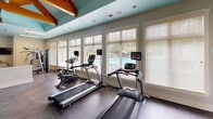 This fitness center is designed to offer a well-rounded workout experience within a bright, thoughtfully appointed space. The room's most distinctive architectural feature is its exposed natural wood truss system, which rises dramatically against a boldly painted teal ceiling, lending a warm yet energetic character to the interior. Neutral gray walls and durable rubber flooring provide a practical, clean backdrop that complements the overall design.

Natural light is a prominent feature, with large windows fitted with white plantation-style blinds lining multiple walls. Several of these windows overlook what appears to be a pool area, providing an engaging outdoor view during workouts. Ceiling fans integrated into the truss structure ensure comfortable air circulation throughout.

The cardio zone is well-stocked with multiple treadmills, elliptical trainers, and stationary bikes, each equipped with individual monitors. A dedicated strength training area features a variety of selectorized cable and resistance machines, along with benches and free-weight equipment. A wall-mounted flat-screen television and a louvered storage door add functional convenience. Teal-painted built-in cubbies near the entrance provide organized storage, rounding out this comprehensively equipped fitness facility.