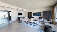 This single-floor space presents a refined and contemporary design, characterized by clean lines, a neutral color palette, and thoughtfully curated furnishings. The main living area is anchored by polished concrete floors that lend an industrial-chic quality to the space, complemented by a plush blue abstract area rug that adds warmth and visual interest. Seating is arranged around a bold rectangular coffee table with a matte black frame, paired with a generous sofa and matching accent chairs upholstered in crisp white fabric. A wall-mounted flat-screen television serves as a focal point on one end of the room, flanked by a compact workspace with a dark-toned desk and chair — ideal for productivity. A striking dark wood wardrobe or cabinet with a gold accent handle introduces a touch of elegance and contrast against the bright white walls. Large-scale abstract artwork in deep blues and charcoals adds a dramatic visual statement to the space. Recessed lighting throughout the ceiling provides even, ambient illumination, enhancing the bright and airy atmosphere. The overall ambiance is modern, polished, and inviting — a well-appointed space suited for both relaxation and function.