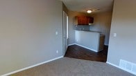 This single-floor space offers a practical and cohesive layout that integrates the living area and kitchen in an open-concept design. The living area is finished with plush neutral carpeting, creating a comfortable and inviting atmosphere, while the kitchen and adjoining spaces transition to rich, dark hardwood-style flooring that adds warmth and contrast.

The kitchen features warm-toned wood cabinetry mounted above a peninsula-style counter, which provides both workspace and a natural divider between the living and kitchen areas. A built-in range or ventilation unit is visible beneath the upper cabinets, pointing to a functional and efficient cooking setup. The countertops offer ample prep space, and the layout supports ease of movement throughout.

The walls are finished in a neutral gray tone throughout, lending the space a clean, modern aesthetic that complements the natural wood tones of the cabinetry and flooring. White baseboards provide a crisp finishing touch. A doorway is positioned along one wall, offering access to additional areas of the unit. Overhead lighting ensures the space is well-lit. Overall, this unit presents a straightforward, well-maintained layout suited for everyday living.