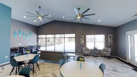 This single-floor communal space is thoughtfully designed to offer both comfort and style. The room immediately draws the eye upward to its vaulted ceilings, which are fitted with two large, statement ceiling fans that provide both visual impact and practical airflow throughout the space. Recessed lighting complements the abundant natural light that pours in through expansive floor-to-ceiling windows, offering unobstructed views of the outdoor pool area beyond.

The interior is finished with sleek marble-patterned tile flooring that lends a clean, upscale feel underfoot. The walls are painted in a sophisticated deep gray tone, creating a modern and welcoming ambiance. Furnishings are tastefully curated, featuring a plush gray sofa and coffee table as a central seating arrangement, flanked by a pair of classic patterned accent chairs positioned near the windows. Teal-accented dining chairs and round tables are arranged throughout the space, adding a vibrant pop of color that ties the design together. Subtle decorative touches, including wall art and fresh floral accents, complete the polished aesthetic. The overall atmosphere is open, airy, and well-suited for socializing or relaxing in a stylish, contemporary setting.