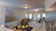 This single-floor residence offers a welcoming and well-appointed interior designed for comfortable everyday living. The open-concept living and dining area serves as the heart of the home, bathed in natural light streaming through multiple windows fitted with blinds and a set of classic French doors that provide both brightness and direct outdoor access. The living space is thoughtfully arranged with plush seating, including a cushioned armchair draped with a cozy throw, a patterned area rug, and a round accent table, creating a relaxed and inviting atmosphere. A ceiling fan overhead adds both function and character to the room. The dining area flows seamlessly from the living space and features a substantial dining table surrounded by elegantly upholstered chairs, positioned to take full advantage of the natural light from the adjacent windows. Neutral wall tones and light-colored carpeting throughout lend the interior a clean, airy quality, while decorative accents and carefully selected furnishings add warmth and personality. A staircase railing visible along one wall hints at architectural detail, contributing to the overall charm of the space. The cohesive design and abundance of natural light make this a bright and functional single-floor home.