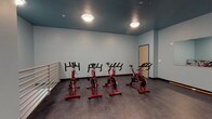 This single-floor indoor cycling studio is purpose-built for group fitness and spin training. The room is finished with cool light-blue walls that contrast sharply with the deep teal-painted ceiling, creating a focused and energizing atmosphere. The dark, durable flooring runs throughout, providing a stable surface suited for high-intensity workouts.

The studio is equipped with several red stationary spin bikes arranged to allow ample space between riders. A large wall mirror spans one side of the room, enabling participants and instructors to monitor form and technique during sessions. A horizontal ballet barre is mounted along one wall, offering supplementary support for stretching and balance exercises.

Exposed metal ductwork runs along the upper portion of the space, contributing to an industrial aesthetic while ensuring effective ventilation during workouts. Flush-mount ceiling light fixtures provide consistent, even illumination throughout the room. A dedicated access door with a blue-trimmed frame is positioned along one wall, providing entry and exit to the studio.

The layout offers open floor space beyond the bike setup, allowing the room to accommodate a variety of fitness activities. The combination of functional equipment, mirrored walls, and a bold, modern color palette makes this studio a well-equipped and visually distinctive space for cycling classes and group exercise.