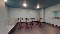 This single-floor indoor cycling studio is purpose-built for group fitness and spin training. The room is finished with cool light-blue walls that contrast sharply with the deep teal-painted ceiling, creating a focused and energizing atmosphere. The dark, durable flooring runs throughout, providing a stable surface suited for high-intensity workouts.

The studio is equipped with several red stationary spin bikes arranged to allow ample space between riders. A large wall mirror spans one side of the room, enabling participants and instructors to monitor form and technique during sessions. A horizontal ballet barre is mounted along one wall, offering supplementary support for stretching and balance exercises.

Exposed metal ductwork runs along the upper portion of the space, contributing to an industrial aesthetic while ensuring effective ventilation during workouts. Flush-mount ceiling light fixtures provide consistent, even illumination throughout the room. A dedicated access door with a blue-trimmed frame is positioned along one wall, providing entry and exit to the studio.

The layout offers open floor space beyond the bike setup, allowing the room to accommodate a variety of fitness activities. The combination of functional equipment, mirrored walls, and a bold, modern color palette makes this studio a well-equipped and visually distinctive space for cycling classes and group exercise.