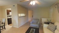 This single-floor space features a comfortable living area with warm, neutral-toned walls and soft carpeted flooring that contribute to a welcoming ambiance. A ceiling fan with an integrated light fixture is centrally mounted, providing both air circulation and overhead lighting. A notable architectural detail is the pass-through window connecting the living area to the kitchen, offering a practical and open feel between the two spaces. The living area is well-proportioned, providing ample room for seating arrangements, as seen with the patterned accent chair and coffee table visible in the space. A hallway leads further into the unit, suggesting additional rooms beyond the main living area. The textured ceiling adds a subtle design element throughout. White baseboards frame the perimeter of the room, providing a clean contrast against the warm wall color. The overall layout and design create a functional and comfortable environment, with a straightforward, practical aesthetic that balances simplicity with warmth.