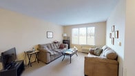 This single-floor residence at Yellowwood offers a practical and comfortable layout across several well-defined living spaces. The living room serves as the central gathering area, finished with soft neutral walls, plush carpeting, and a textured ceiling. A large multi-pane window floods the room with natural light and provides views of the surrounding green outdoor space, creating an open and airy atmosphere. A wall-mounted unit provides in-room climate control.

The bedroom is a quiet, carpeted retreat featuring its own window with outdoor views, a spacious closet with a warm wood-toned door, and ample room for furniture including a full dresser and shelving. The neutral color palette carries throughout, contributing to a calm and cohesive feel.

A compact dining area provides a dedicated space for meals, accommodating a small round table and chairs. It connects naturally to the surrounding living spaces, making the overall layout feel efficient and functional.

The bathroom is designed with accessibility in mind, featuring a roll-in style tiled shower with grab bars, a handheld showerhead, and a medicine cabinet with mirror. The space prioritizes ease of use while maintaining a clean, straightforward finish throughout.