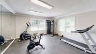 Located at 2421 Chase Park Dr in Montgomery, Alabama, this fitness center offers a well-organized exercise space designed to support a variety of workout routines. The room features durable rubber-mat flooring, crown molding, and a textured ceiling with bright overhead fluorescent lighting that ensures excellent visibility throughout. A full-length wall mirror spans one side of the room, visually expanding the space and providing users with proper form feedback during workouts. Natural light filters in through multiple windows, adding to the bright and energizing atmosphere. The facility is equipped with a diverse selection of exercise equipment, including a treadmill, elliptical trainer, stationary spin bike, and a multi-function cable strength machine, catering to both cardiovascular and resistance training needs. A freestanding punching bag rounds out the equipment offerings for those seeking combat-style fitness training. A wall-mounted flat-screen television provides entertainment during workouts. The space also includes posted gym rules and safety signage, maintaining an organized and user-friendly environment. Access is through a standard door, and the overall layout is efficient and practical, making the most of the available floor area to deliver a comprehensive fitness experience.