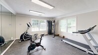 Located at 2421 Chase Park Dr in Montgomery, Alabama, this fitness center offers a well-organized exercise space designed to support a variety of workout routines. The room features durable rubber-mat flooring, crown molding, and a textured ceiling with bright overhead fluorescent lighting that ensures excellent visibility throughout. A full-length wall mirror spans one side of the room, visually expanding the space and providing users with proper form feedback during workouts. Natural light filters in through multiple windows, adding to the bright and energizing atmosphere. The facility is equipped with a diverse selection of exercise equipment, including a treadmill, elliptical trainer, stationary spin bike, and a multi-function cable strength machine, catering to both cardiovascular and resistance training needs. A freestanding punching bag rounds out the equipment offerings for those seeking combat-style fitness training. A wall-mounted flat-screen television provides entertainment during workouts. The space also includes posted gym rules and safety signage, maintaining an organized and user-friendly environment. Access is through a standard door, and the overall layout is efficient and practical, making the most of the available floor area to deliver a comprehensive fitness experience.