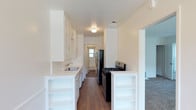 This single-floor residence offers a clean, functional layout with thoughtfully designed living spaces. The kitchen serves as a central highlight of the home, designed in a classic galley style that maximizes efficiency and storage. White shaker-style cabinetry lines both sides of the kitchen, extending from floor to ceiling to provide ample storage space. The countertops are finished in a crisp white surface, complementing the bright, airy aesthetic of the room. A stainless steel sink is neatly integrated into the cabinetry, while black appliances — including a gas range and a stainless steel refrigerator — add a striking contrast to the otherwise light color palette. Open shelving built into the lower cabinetry offers additional display and storage versatility. Rich hardwood flooring runs the length of the kitchen, lending warmth and character to the space. Natural light filters in through a window at the far end of the kitchen, enhancing the sense of openness. Adjacent to the kitchen, a carpeted living area is visible, providing a comfortable transition between spaces. The overall ambiance of the home is fresh, clean, and move-in ready, with a neutral palette that offers flexibility for personalization.