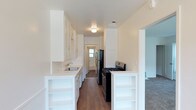 This single-floor residence offers a clean, functional layout with thoughtfully designed living spaces. The kitchen serves as a central highlight of the home, designed in a classic galley style that maximizes efficiency and storage. White shaker-style cabinetry lines both sides of the kitchen, extending from floor to ceiling to provide ample storage space. The countertops are finished in a crisp white surface, complementing the bright, airy aesthetic of the room. A stainless steel sink is neatly integrated into the cabinetry, while black appliances — including a gas range and a stainless steel refrigerator — add a striking contrast to the otherwise light color palette. Open shelving built into the lower cabinetry offers additional display and storage versatility. Rich hardwood flooring runs the length of the kitchen, lending warmth and character to the space. Natural light filters in through a window at the far end of the kitchen, enhancing the sense of openness. Adjacent to the kitchen, a carpeted living area is visible, providing a comfortable transition between spaces. The overall ambiance of the home is fresh, clean, and move-in ready, with a neutral palette that offers flexibility for personalization.