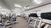 Located at 6795 Belle Vale Dr in Addis, Louisiana, this fitness center offers a generously sized, single-floor exercise room designed to support a full range of workout routines. The space is laid out in a long, open rectangular floor plan with carpet flooring throughout, providing a comfortable and functional environment for exercise.

The room is well-lit with recessed fluorescent lighting and complemented by dual ceiling fans that promote air circulation. Multiple windows fitted with horizontal blinds line the walls, allowing natural light to filter in and creating a bright, inviting atmosphere. Full-length mirrors along one wall enhance the sense of space and allow users to monitor their form during workouts.

The facility is comprehensively equipped with a diverse selection of fitness machines and free weights. Cardio equipment includes treadmills, elliptical machines, and stationary cycling bikes. Strength training is well represented with a variety of selectorized cable machines and multi-function weight stations. A dedicated free weight area features a full dumbbell rack with a flat bench for targeted exercises. A body weight scale is also available near the exit. Crown molding along the ceiling adds a refined finishing touch to the space, elevating the overall aesthetic beyond a typical utilitarian gym setting.