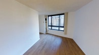 This single-floor space offers a clean, functional layout with a bright and open feel. The main living area is highlighted by warm hardwood-style flooring that runs throughout, complementing the crisp white walls and creating a welcoming, neutral aesthetic. A large picture window with a black frame serves as a standout architectural feature, allowing abundant natural light to pour into the space while offering views of the surrounding urban exterior. Sheer cream-colored curtains frame the window, adding a soft touch to the otherwise streamlined interior. The textured ceiling adds subtle visual interest overhead, while clean baseboards and smooth wall finishes contribute to the overall well-maintained condition of the space. A wall-mounted control panel is conveniently positioned near the window, suggesting integrated climate or intercom functionality. The open floor plan provides flexibility for a variety of furniture arrangements, making efficient use of the available area. The overall ambiance is light, airy, and move-in ready, with a minimalist character that offers a versatile canvas for personalization.