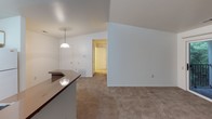 This single-floor unit is thoughtfully laid out to maximize its compact footprint, offering one bedroom and one bathroom across a well-organized floor plan. The living area features vaulted ceilings that add a sense of openness, neutral-toned carpet flooring, and a sliding glass door leading to a private balcony framed by lush green views. A pendant light fixture hangs near the kitchen peninsula, providing both task lighting and a subtle visual anchor between the living and kitchen spaces.

The kitchen is efficiently arranged in an L-shaped configuration, featuring white upper cabinetry, a dark laminate countertop that wraps around the peninsula, a double-basin sink, a white electric range, an over-the-range microwave, and a full-size refrigerator. Wood-look flooring runs through the kitchen, transitioning to carpet in the adjoining areas.

The bedroom is a generously sized room with two windows fitted with blinds, allowing for natural light and views of surrounding greenery. Carpet continues throughout. The bathroom includes wood-look flooring, a single-sink vanity with under-cabinet storage, a wall-mounted mirror, and a bathtub/shower combination.