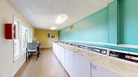 Located at 189 Wallace Rd in Nashville, Tennessee, this community laundry facility offers a well-organized and functional space designed to serve residents efficiently. The single-floor layout is thoughtfully arranged, with a long row of top-load washing machines lining the teal-painted accent wall, complemented by a bank of stacked dryer units positioned along the opposite end of the room. The light tile flooring runs throughout, making the space easy to maintain and navigate. Overhead fluorescent lighting ensures the room is well-lit at all times. A textured ceiling adds a classic touch to the interior. At one end of the facility, a bright and welcoming seating nook features large windows that allow ample natural light to filter in, offering outdoor views of the surrounding greenery. This corner includes a table and chairs, providing a comfortable spot for residents to wait. A wall-mounted folding shelf offers additional convenience for sorting laundry. Informational bulletin boards and posted notices keep residents informed, while a fire safety panel mounted on the wall reflects the facility's attention to safety standards. The warm yellow tones of the waiting area contrast pleasantly with the cool teal walls of the main laundry area, creating a clean and inviting atmosphere throughout.