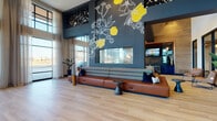 This contemporary communal space makes an immediate impression with its soaring double-height ceilings and a dramatic navy accent wall adorned with a hand-illustrated floral mural in bold yellow and white. A sleek linear fireplace is centered within the mural wall, flanked by a generously scaled cognac leather banquette with layered throw pillows, creating a warm and inviting focal point for the main lounge area. Light wood plank flooring extends throughout, lending warmth against the cool, dark architectural palette of navy, charcoal, and black metal accents.

Floor-to-ceiling windows and sheer curtains line the perimeter, flooding the interior with abundant natural light and offering open views to the outdoors. A pair of cognac leather lounge chairs positioned near the expansive glazing provides an intimate seating vignette perfect for relaxed conversation or quiet reflection.

Beyond the main lounge, the space transitions into a dining and social zone featuring semi-private booth seating upholstered in deep green velvet, complemented by patterned carpet and pendant lighting. A bar and kitchenette area with navy cabinetry, stone backsplash, and a wine refrigerator adds a refined hospitality element. Decorative fluted columns and a corridor lined with glass-walled private rooms complete the layout, offering flexible spaces for focused work or small gatherings.