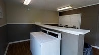 This single-floor communal laundry facility is designed with practicality and efficiency in mind. The room features multiple top-loading washing machines and a row of dryers, providing ample capacity to accommodate shared use. A continuous white folding counter runs along the center of the space, offering a convenient surface for sorting and folding laundry. The room's two-tone walls — with a darker lower panel and lighter upper section accented by a brown border — give the space a clean, structured appearance. Durable dark wood-style flooring covers the ground, providing a low-maintenance surface well-suited to a high-traffic environment. Overhead fluorescent lighting ensures the space is bright and well-illuminated throughout. The textured ceiling adds a subtle design element while contributing to the room's functional aesthetic. Overall, this laundry facility is thoughtfully arranged to maximize usability, offering residents a well-equipped and organized space for their laundry needs.