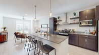 This single-floor residence showcases a thoughtfully designed open-concept layout where the kitchen and living area flow seamlessly together. The kitchen is a standout feature, equipped with dark espresso cabinetry, a stainless steel appliance suite including a range with an electric cooktop, a full-size refrigerator, and a built-in microwave. A stainless wall-mounted range hood serves as a sleek focal point, while open floating shelves add a modern, airy touch. The large central island, topped with a neutral quartz-style countertop, provides ample prep space and accommodates bar seating beneath stylish glass pendant lights. Light wood-toned flooring runs throughout, complementing the bright, white walls.

The living area receives generous natural light through French doors, which also provide outdoor access. A ceiling fan overhead adds comfort to the space. The bedroom is a calm retreat with a window allowing natural light, a ceiling fan, and light wood flooring. The bathroom features a tub-shower combination with clean white tile, dark cabinetry with matching countertops, and brushed nickel fixtures. A secondary bathroom area includes in-unit laundry facilities, adding everyday convenience.