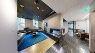 Located at 1 India St in Boston's historic waterfront neighborhood, this thoughtfully designed single-floor space offers a refined blend of modern aesthetics and functional versatility. The layout is organized into distinct yet interconnected zones, each with its own character and purpose.

The lounge area serves as a striking focal point, anchored by a dramatic floor-to-ceiling marble fireplace with an open flame insert and a wall-mounted display above. Stylish lounge seating is arranged atop a rich blue area rug, complemented by a textured dark acoustic ceiling with recessed and track lighting that creates a warm, intimate atmosphere. Natural light filters in through street-facing windows.

Transitioning seamlessly from the lounge, the open dining and collaboration area features a large wood-topped communal table with bar-height seating, illuminated by a trio of pendant lights. Light hardwood flooring contrasts with the hexagonal tile that grounds the central corridor, adding visual interest and defining circulation paths.

Steel-framed glass partitions enclose a series of private meeting rooms, maintaining an open, airy feel while providing acoustic separation. One room accommodates a full conference table with colorful seating, while a smaller nook offers an intimate round table setting. Throughout, curated artwork adorns the walls, enhancing the polished, professional ambiance of this well-appointed Boston space.