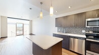 Located at 1028 N Hamilton St in Spokane, WA, this modern 2-bedroom, 1-bathroom unit offers a well-designed open-concept layout with contemporary finishes throughout. The kitchen serves as a central highlight, featuring dark espresso cabinetry, a mosaic tile backsplash, sleek white quartz countertops, a center island with pendant lighting, and a full suite of stainless steel appliances including a dishwasher, range, microwave, and refrigerator. The kitchen flows naturally into the living area, which is bathed in natural light from large floor-to-ceiling windows and a glass balcony door that opens to a private outdoor balcony with open views of the surrounding area. Warm hardwood-style flooring runs throughout the main living spaces and bedrooms, adding a cohesive and inviting feel. The spacious primary bedroom receives abundant natural light and offers direct balcony access. A dedicated walk-in closet features built-in wire shelving for organized storage. The full bathroom includes a tub-shower combination with brushed nickel fixtures and tile flooring. A hallway connects the private rooms, maintaining a clean separation between living and sleeping areas. The unit also includes in-unit laundry, as seen in the floorplan overview.