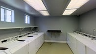This shared laundry facility is designed with practicality and efficiency in mind. The room features a well-organized layout with a row of top-loading washing machines and matching dryers arranged along two walls, maximizing the available floor space and allowing multiple users to do laundry simultaneously. A wall-mounted utility shelf on one side provides convenient storage, while a dark-topped folding table anchored to the opposite wall offers a dedicated surface for sorting and folding clothes. The walls are finished in a clean, neutral grey tone that contributes to the room's tidy and orderly atmosphere. Overhead, large recessed fluorescent panel lights illuminate the entire space evenly, ensuring excellent visibility throughout. A small window on one wall allows a modest amount of natural light to supplement the artificial lighting. The flooring is a durable, easy-to-clean surface well-suited to the demands of a high-use laundry environment. Overall, this laundry room is a well-equipped, no-frills facility that prioritizes functionality, cleanliness, and convenience for its users.