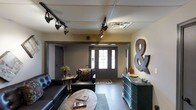 This single-floor space exudes an industrial-chic aesthetic, blending raw character with carefully curated design elements. The main lounge area serves as the heart of the space, anchored by dark leather sofas and a rustic round coffee table that adds warmth and texture to the room. A teal vintage-style dresser along the wall provides both storage and a striking decorative focal point, complemented by an oversized metal ampersand wall piece that adds a bold, artistic touch.

The ceiling features a grid-tile design lined with adjustable track lighting, allowing for flexible, directed illumination throughout the space. The dark hardwood flooring grounds the room with a sense of depth and continuity. Neutral walls in muted tones create a balanced backdrop that lets the curated furnishings stand out.

Natural light filters in through a set of classic French doors at the far end of the room, framed by dark-painted trim that reinforces the moody, sophisticated atmosphere. The doorway leads to an adjacent area, hinting at additional functional space beyond. A small side table with a potted plant introduces a natural element, softening the industrial feel. Overall, the space combines eclectic décor, purposeful lighting, and a cohesive dark-toned palette to create an inviting yet visually dynamic environment.