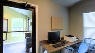 This single-floor space features a functional workspace centered around a built-in desk with a granite-style countertop surface, accommodating a desktop computer monitor, keyboard, and printer. A corkboard panel mounted on the neutral gray wall above the desk provides a practical organizational surface. The workspace is compact yet efficient, offering dedicated areas for computing and administrative tasks.

A dark-framed glass door connects this workspace to an adjacent room, which is visible through the opening. That neighboring space features vaulted ceilings, yellow-green accent walls, and large windows that allow generous natural light to filter through, adding a sense of openness to the overall environment. Dark cabinetry and fixtures are visible in the adjoining room, suggesting a utility or grooming area. A fire extinguisher mounted near the doorway indicates attention to safety protocols, pointing to a professional or commercial use for the space. The overall layout is practical and organized, designed to support productivity within a compact footprint.
