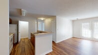 This single-story residence is designed with a functional, open-concept layout that connects the kitchen and living areas into one cohesive space. The kitchen is positioned around a peninsula counter that doubles as a breakfast bar, offering a natural boundary between the cooking and living zones. Warm-toned wood cabinetry provides ample storage, while white appliances — including a coil-burner electric range with overhead ventilation and a refrigerator — deliver practical utility. Neutral countertops complement the overall palette throughout.

The living area is generously proportioned, featuring warm hardwood-style flooring that continues from the kitchen. A sliding glass door at one end of the living space allows natural light to fill the room and provides outdoor access. Textured ceilings are present throughout the main living and kitchen areas, adding subtle visual interest overhead.

Two bedrooms are fitted with plush carpet and individual windows that allow natural light in. Each room offers a clean, neutral finish suitable for a range of furnishing configurations.

The bathroom includes a vanity with wood cabinetry and a large mirror, a tub-shower combination surrounded by white square tile, hardwood-style flooring, and standard fixtures — providing a complete, well-organized space.