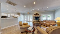 Located at 2162 Millstream Cir in Creedmoor, NC, this single-story community building offers a welcoming and well-appointed interior designed for comfort and functionality. The main lounge area is generously sized, showcasing light hardwood flooring, soft neutral wall tones, and elegant crown molding throughout. A standout feature is the floor-to-ceiling river rock fireplace with a rustic wood mantel, serving as a dramatic focal point for the seating area, which is furnished with leather sofas and armchairs arranged around glass-topped coffee tables. Recessed lighting and a ceiling fan provide practical illumination and airflow, while multiple sets of French doors and large windows flood the space with natural light and offer views of the surrounding greenery. A reception or office area is positioned toward the front of the building, complete with a built-in desk workspace and additional seating. The kitchen is fully equipped with white cabinetry, a full-size refrigerator, electric range, microwave, dishwasher, and generous counter space, along with a peninsula that separates it from the main gathering area. The space is thoughtfully designed to accommodate both casual social gatherings and organized community functions, making it a versatile and inviting hub for residents.