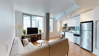 This single-floor space showcases a thoughtfully designed open-concept layout that seamlessly integrates the living and kitchen areas into one cohesive environment. Large floor-to-ceiling windows line the living area, flooding the space with abundant natural light while framing elevated views of the surrounding urban skyline. The windows create a bright, airy atmosphere that enhances the overall sense of openness.

A distinctive concrete column serves as a striking architectural feature, adding an industrial character that contrasts elegantly with the otherwise contemporary aesthetic. Warm-toned hardwood flooring flows continuously throughout, grounding the space and contributing to its inviting feel.

The kitchen is positioned along one wall and features sleek, high-gloss white cabinetry, stainless steel appliances including a built-in microwave and range, and dark granite countertops. Track lighting above the kitchen provides focused illumination, complementing the natural light from the nearby windows.

The living area accommodates a tufted sofa, media console, and a round coffee table arranged in a relaxed, functional configuration. Subtle greenery accents the space, softening the clean, modern lines throughout.