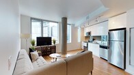 This single-floor space showcases a thoughtfully designed open-concept layout that seamlessly integrates the living and kitchen areas into one cohesive environment. Large floor-to-ceiling windows line the living area, flooding the space with abundant natural light while framing elevated views of the surrounding urban skyline. The windows create a bright, airy atmosphere that enhances the overall sense of openness.

A distinctive concrete column serves as a striking architectural feature, adding an industrial character that contrasts elegantly with the otherwise contemporary aesthetic. Warm-toned hardwood flooring flows continuously throughout, grounding the space and contributing to its inviting feel.

The kitchen is positioned along one wall and features sleek, high-gloss white cabinetry, stainless steel appliances including a built-in microwave and range, and dark granite countertops. Track lighting above the kitchen provides focused illumination, complementing the natural light from the nearby windows.

The living area accommodates a tufted sofa, media console, and a round coffee table arranged in a relaxed, functional configuration. Subtle greenery accents the space, softening the clean, modern lines throughout.