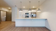 Located at 7950 Park Blvd N in Pinellas Park, Florida, this contemporary single-floor residence offers a clean, modern aesthetic with thoughtfully designed living spaces. The open-concept layout seamlessly connects the kitchen and living area, creating a bright and airy atmosphere enhanced by recessed lighting and neutral-toned walls throughout.

The kitchen is a standout feature, equipped with stainless steel appliances including a range, microwave, and French door refrigerator. Crisp white shaker-style cabinetry extends to the ceiling, providing generous storage, while a stone-textured tile backsplash adds visual interest. A large granite-topped island serves as the centerpiece, offering additional prep space and seating. Three industrial-style chrome pendant lights hang above the island, lending a modern, polished touch.

The living area flows naturally from the kitchen, featuring light wood-style plank flooring that carries a warm, cohesive feel across the main living spaces. The bedroom is carpeted for comfort and includes a window that brings in natural light. The bathroom is well-appointed with a granite vanity, white cabinetry, and a tub-shower combination with clean white tile surround. A two-panel door in a muted gray finish adds a contemporary design detail. Overall, the space delivers a fresh, move-in-ready environment with consistent modern finishes.
