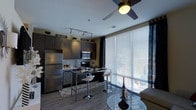 This single-floor unit showcases a thoughtfully designed open-concept layout that seamlessly integrates the kitchen, dining, and living areas. The kitchen is outfitted with dark espresso cabinetry, stainless steel appliances including a full-size refrigerator, a built-in microwave, and a range with overhead ventilation. A track lighting system runs along the ceiling, providing focused illumination throughout the cooking space. A compact kitchen island with bar-height seating serves as a functional divider between the kitchen and the combined dining and living area. Light wood-tone flooring extends throughout the space, creating a clean, contemporary aesthetic that contrasts with the rich, dark cabinetry. A ceiling fan with an integrated light fixture adds both comfort and a modern touch overhead. Floor-to-ceiling windows dressed with horizontal blinds and dark curtain panels flood the space with natural light while offering privacy control. The living area accommodates a seating arrangement alongside a glass-topped accent table, contributing to the unit's sleek, modern ambiance overall.