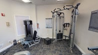 Located at 3232 N Locust St in Denton, Texas, this fitness center is a dedicated, single-floor exercise facility designed to support a comprehensive workout routine. The space features durable patterned carpet flooring throughout and is finished with clean white walls, creating a bright and functional environment. Overhead fluorescent lighting ensures the room is well-illuminated, while a ceiling fan adds to the comfort of the workout area.

A standout feature of the main exercise area is the full-wall mirror, which expands the sense of space and allows users to monitor their form during exercise. The room is entered through elegant French doors, lending a refined touch to an otherwise utilitarian setting.

The facility is equipped with a well-rounded selection of Life Fitness strength machines, including a leg press and a multi-function cable/functional trainer station. Cardio options include a treadmill and an elliptical trainer. A dedicated free weight zone offers a dumbbell rack with multiple weight increments. An exercise guide chart is posted on the wall for user reference, and a clearly displayed Fitness Center Rules sign promotes safe usage of the space.

Natural light filters in through several windows with horizontal blinds, offering views of the surrounding greenery. Safety equipment, including a fire extinguisher and fire alarm, is also present throughout the space.