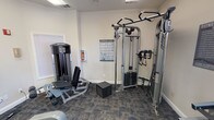 Located at 3232 N Locust St in Denton, Texas, this fitness center is a dedicated, single-floor exercise facility designed to support a comprehensive workout routine. The space features durable patterned carpet flooring throughout and is finished with clean white walls, creating a bright and functional environment. Overhead fluorescent lighting ensures the room is well-illuminated, while a ceiling fan adds to the comfort of the workout area.

A standout feature of the main exercise area is the full-wall mirror, which expands the sense of space and allows users to monitor their form during exercise. The room is entered through elegant French doors, lending a refined touch to an otherwise utilitarian setting.

The facility is equipped with a well-rounded selection of Life Fitness strength machines, including a leg press and a multi-function cable/functional trainer station. Cardio options include a treadmill and an elliptical trainer. A dedicated free weight zone offers a dumbbell rack with multiple weight increments. An exercise guide chart is posted on the wall for user reference, and a clearly displayed Fitness Center Rules sign promotes safe usage of the space.

Natural light filters in through several windows with horizontal blinds, offering views of the surrounding greenery. Safety equipment, including a fire extinguisher and fire alarm, is also present throughout the space.