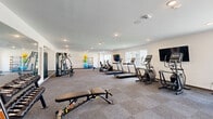 Located at 14744 Malachi Rd in Noblesville, Indiana, this well-appointed fitness center offers a generous, open layout designed to support a wide range of workout routines. The space is finished with neutral-toned carpeting throughout and smooth white walls accented by recessed lighting, creating a clean and motivating environment.

The cardio zone is well-stocked, featuring multiple treadmills, elliptical trainers, a stationary bike, and a rowing machine, all arranged in an orderly row along the main floor. Two wall-mounted flat-screen TVs are positioned for easy viewing during workouts. A dedicated strength training area includes a multi-function cable machine, a dumbbell rack with an adjustable weight bench, and a full-length wall mirror that enhances the sense of space and allows for proper form checks.

A freestanding rack along one wall holds an assortment of exercise balls and weighted accessories, supporting stretching and functional training exercises. Multiple windows line the walls, inviting natural light and providing views of the surrounding community. The open floor plan allows for ample circulation between equipment zones, making the space practical and accessible for users of all fitness levels.