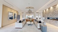 This expansive single-floor amenity space at Hanover Collins Place is a showcase of contemporary design and refined elegance. The welcoming lobby lounge serves as the heart of the space, featuring a striking geometric pendant chandelier suspended from a circular ceiling detail, flanked by white columns and warm wood accent walls. Two plush neutral sofas frame a round marble-top coffee table atop a patterned area rug, creating an inviting seating arrangement. Abstract artwork adorns the walls, contributing to an elevated, gallery-like atmosphere. A textured herringbone stone feature wall with a built-in linear fireplace anchors one side of the lounge, while floor-to-ceiling windows at the rear flood the interior with natural light and offer views of the outdoor amenity terrace complete with grilling stations. The communal kitchen is a standout feature, centered around a generous waterfall-edge quartz island with bar seating beneath dramatic black cone pendant lights. A golden herringbone-paneled tray ceiling overhead adds warmth and sophistication. Light herringbone-patterned flooring flows seamlessly throughout the space. A media lounge area with a built-in entertainment wall and custom cabinetry completes this thoughtfully designed communal environment.