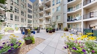 This thoughtfully designed outdoor courtyard serves as a central gathering space, fully enclosed by a modern multi-story residential building featuring horizontal lap siding, large windows, and private balconies on every floor. The courtyard is organized into several distinct seating zones, each offering a unique atmosphere for relaxation and socializing. A standout fire pit seating area anchors one section of the space, surrounded by contemporary lounge chairs and accent stools, ideal for gathering in the evenings. Overhead string lights extend across the courtyard, adding a warm, inviting ambiance after dark. Another seating vignette features sleek high-back chairs arranged around a bistro-style table, providing a more intimate setting. The hardscape is composed of large-format concrete pavers laid in a clean grid pattern, while gravel beds and generously sized planters introduce rich layers of greenery and seasonal color. Vibrant flowering plants — including petunias, black-eyed Susans, and ornamental grasses — bring life and vibrancy to the space throughout the warmer months. A ground-level railed terrace along one edge of the building offers additional outdoor seating. The open-sky design allows for abundant natural light, making this courtyard a welcoming and functional outdoor retreat.