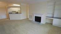 This single-floor residence offers a well-organized and inviting layout centered around a spacious, open living area. The room is finished with neutral wall tones and soft wall-to-wall carpeting, creating a warm and understated ambiance throughout. A standout architectural feature is the classic white fireplace, framed by elegant columned millwork and a traditional mantel surround, which serves as a natural focal point of the main living space. Crown molding trims the ceiling, adding a refined touch to the overall design. Adjacent to the living area, the kitchen is connected via a pass-through counter that promotes an open feel between the two spaces. The kitchen is equipped with white cabinetry, a refrigerator, and a sink beneath overhead lighting, offering a practical and bright cooking environment. A wall-mounted HVAC vent ensures year-round climate comfort. The neutral color palette, consistent flooring, and clean architectural lines throughout contribute to a cohesive and move-in-ready living environment that is both functional and comfortable.