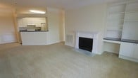 This single-floor residence offers a well-organized and inviting layout centered around a spacious, open living area. The room is finished with neutral wall tones and soft wall-to-wall carpeting, creating a warm and understated ambiance throughout. A standout architectural feature is the classic white fireplace, framed by elegant columned millwork and a traditional mantel surround, which serves as a natural focal point of the main living space. Crown molding trims the ceiling, adding a refined touch to the overall design. Adjacent to the living area, the kitchen is connected via a pass-through counter that promotes an open feel between the two spaces. The kitchen is equipped with white cabinetry, a refrigerator, and a sink beneath overhead lighting, offering a practical and bright cooking environment. A wall-mounted HVAC vent ensures year-round climate comfort. The neutral color palette, consistent flooring, and clean architectural lines throughout contribute to a cohesive and move-in-ready living environment that is both functional and comfortable.