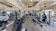 Located at 1475 Benton Blvd in Pooler, Georgia, this single-story fitness center offers a comprehensive exercise environment designed for a full range of workout routines. The main exercise area is generously proportioned and outfitted with an extensive selection of professional-grade equipment, including Cybex and True branded strength machines, treadmills, ellipticals, recumbent bikes, and upright cycles. Patterned carpet flooring runs throughout the main floor, providing a durable and practical surface suited to high-traffic use.

The architecture features elegant coffered ceilings with recessed lighting and multiple ceiling fans, ensuring the space remains well-lit and comfortable. Large windows line the perimeter walls, allowing abundant natural light to pour in while offering views of the surrounding outdoor greenery. Floor-to-ceiling mirrors are strategically positioned throughout the main workout area, visually expanding the space and allowing users to monitor their form during exercise.

A dedicated studio room is also available, finished with warm hardwood flooring, soft sage-green walls, a wall-mounted mirror with ballet barre, and recessed lighting. This room is equipped with yoga mats, a stability ball, and a step platform, making it well-suited for stretching, yoga, or group fitness activities. An entry corridor connects the spaces and features additional windows and posted facility guidelines.