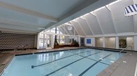This impressive indoor aquatic facility is defined by its striking architectural design, centered around a pristine lap pool with clearly marked lanes. The space is anchored by a dramatic vaulted ceiling with exposed white-painted rafters that rise to a generous apex, creating an airy, open atmosphere that feels expansive and inviting.

Natural light floods the interior through a generous wall of floor-to-ceiling windows and glass panels along one side, offering views to the outdoors while maintaining a bright, welcoming environment year-round. A skylight integrated into the upper portion of the roofline further enhances the sense of openness.

Along the poolside, a warm wood-toned bench and planter area provides a comfortable lounge space, accented with colorful cushions and lush greenery, adding a touch of comfort and vitality to the setting. Bold chevron-patterned wall décor on one end adds a modern, stylish contrast to the clean white structural elements.

The pool enclosure includes access doors leading to adjacent areas, and informational signage is neatly mounted on the walls. Mirrored panels on one end of the facility reflect light and contribute to the feeling of spaciousness. The overall ambiance is clean, bright, and functional, making this an exceptional space for both recreational swimming and structured aquatic programming.