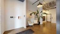 This single-floor lobby showcases a thoughtfully designed industrial-modern aesthetic that sets a distinctive tone upon entry. The space features polished concrete floors that reflect light and contribute to the open, airy feel of the interior. Soaring ceilings with fully exposed ductwork, conduit, and structural elements lend an authentic industrial character, while matte black pendant lights provide focused, warm illumination throughout.

Structural columns with a clean white finish anchor the space visually, complementing the crisp white walls that serve as a gallery-like backdrop. Curated wall-hung textile artwork adds an artistic dimension, while a pair of classic white Barcelona-style chairs arranged around the columns creates a refined and functional seating area. A large bird-of-paradise plant introduces vibrant greenery, softening the industrial elements.

A bank of built-in mailboxes lines one wall, offering a practical residential or mixed-use function. The lobby also features a stainless steel elevator and a secondary access door, ensuring efficient circulation. Large glass entry doors allow natural light to filter in, brightening the polished floor and reinforcing the space's clean, open character. Overall, the lobby balances industrial architecture with contemporary furnishings to create a welcoming and visually cohesive environment.