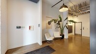 This single-floor lobby showcases a thoughtfully designed industrial-modern aesthetic that sets a distinctive tone upon entry. The space features polished concrete floors that reflect light and contribute to the open, airy feel of the interior. Soaring ceilings with fully exposed ductwork, conduit, and structural elements lend an authentic industrial character, while matte black pendant lights provide focused, warm illumination throughout.

Structural columns with a clean white finish anchor the space visually, complementing the crisp white walls that serve as a gallery-like backdrop. Curated wall-hung textile artwork adds an artistic dimension, while a pair of classic white Barcelona-style chairs arranged around the columns creates a refined and functional seating area. A large bird-of-paradise plant introduces vibrant greenery, softening the industrial elements.

A bank of built-in mailboxes lines one wall, offering a practical residential or mixed-use function. The lobby also features a stainless steel elevator and a secondary access door, ensuring efficient circulation. Large glass entry doors allow natural light to filter in, brightening the polished floor and reinforcing the space's clean, open character. Overall, the lobby balances industrial architecture with contemporary furnishings to create a welcoming and visually cohesive environment.
