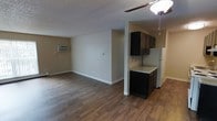 This single-floor space features an open-concept layout that seamlessly connects the living area and kitchen, creating a functional and cohesive environment. The rich, dark wood-style laminate flooring extends throughout, providing a warm and consistent aesthetic across the space.

The living area is spacious and versatile, offering ample room for various furniture arrangements. A ceiling fan with a light fixture provides overhead illumination and air circulation, while a wall-mounted air conditioning unit ensures climate comfort. The neutral beige walls and white trim contribute to a clean, understated look, and a textured popcorn ceiling adds a classic touch.

The kitchen is partially separated by a structural column, lending a sense of definition without fully closing off the space. Dark espresso-toned cabinetry provides bold contrast against the light granite-style countertop. A white refrigerator anchors the kitchen area, and the layout appears compact yet efficient. The transition between the kitchen's flooring and the main living area is smooth and cohesive. Overall, the space presents a straightforward, practical design with contrasting dark and light elements throughout.