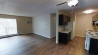 This single-floor space features an open-concept layout that seamlessly connects the living area and kitchen, creating a functional and cohesive environment. The rich, dark wood-style laminate flooring extends throughout, providing a warm and consistent aesthetic across the space.

The living area is spacious and versatile, offering ample room for various furniture arrangements. A ceiling fan with a light fixture provides overhead illumination and air circulation, while a wall-mounted air conditioning unit ensures climate comfort. The neutral beige walls and white trim contribute to a clean, understated look, and a textured popcorn ceiling adds a classic touch.

The kitchen is partially separated by a structural column, lending a sense of definition without fully closing off the space. Dark espresso-toned cabinetry provides bold contrast against the light granite-style countertop. A white refrigerator anchors the kitchen area, and the layout appears compact yet efficient. The transition between the kitchen's flooring and the main living area is smooth and cohesive. Overall, the space presents a straightforward, practical design with contrasting dark and light elements throughout.