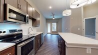 Located at 2001 W Cold Spring in Baltimore, MD, this single-floor residence offers a thoughtfully designed open layout with modern finishes throughout. The kitchen is a standout feature, boasting rich dark-toned cabinetry, stainless steel appliances including a range, microwave, dishwasher, and refrigerator, complemented by crisp white countertops and a generous central island with pendant lighting overhead. Dark hardwood-style flooring runs throughout the kitchen and main living areas, adding warmth and visual continuity. A ceiling fan and access to a private balcony through glass-paned doors bring natural light and ventilation into the open living space. The bedroom areas are carpeted in neutral tones, providing a soft and comfortable feel, with large windows that invite ample natural light and offer views of the surrounding exterior. The bathroom is finished with clean white subway tile surrounding a soaking tub and shower combination, accented by brushed nickel fixtures for a polished look. Additional conveniences include an in-unit washer and dryer, a walk-in pantry or storage closet, and recessed lighting throughout. The overall design palette of soft grays, warm browns, and white creates a cohesive, contemporary ambiance throughout the space.