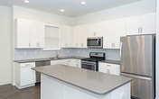 This single-floor space showcases a thoughtfully designed open-concept kitchen that serves as the heart of the layout. The kitchen is outfitted with crisp white shaker-style cabinetry extending to the ceiling, offering generous storage throughout. A neutral gray tile backsplash adds texture and visual interest against the clean white surfaces. Stainless steel appliances, including a French door refrigerator, gas range, and built-in microwave, provide a polished, contemporary look. The expansive central island features a smooth gray quartz-style countertop, offering ample workspace and serving as a natural gathering point. Recessed lighting is installed throughout the ceiling, ensuring bright, even illumination across the space. The warm-toned hardwood-style flooring grounds the room and creates a cohesive contrast to the lighter upper elements. The open sightlines extend beyond the kitchen into an adjoining area, reinforcing the spacious, connected feel of the layout. A doorway visible in the background hints at additional rooms, rounding out the functional and well-appointed nature of this modern space.