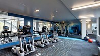 This single-floor fitness center is thoughtfully designed to provide a comprehensive workout environment. The space features a sleek, contemporary aesthetic defined by bold navy blue accent walls that are complemented by large floor-to-ceiling mirrors, creating a sense of expanded space and allowing users to monitor their form during exercise. A nature-inspired mural adds a calming visual element to the strength training area.

The cardio section is well-appointed with a row of modern True-brand ellipticals and additional cardio machines, all positioned to maximize the open floor plan. The strength training area is equipped with a multi-station cable and weight machine system, offering a variety of resistance-based workout options.

Underfoot, a durable patterned carpet with blue accent striping ties the room's color scheme together while providing comfort and traction. Recessed lighting and flush-mounted fluorescent fixtures illuminate the space evenly throughout, ensuring a bright and energizing atmosphere.

The ceiling features an angled design element that adds architectural interest, while a ceiling fan contributes to air circulation during intense workouts. Natural light filters in through windows, further enhancing the open and inviting ambiance of this well-maintained fitness facility.